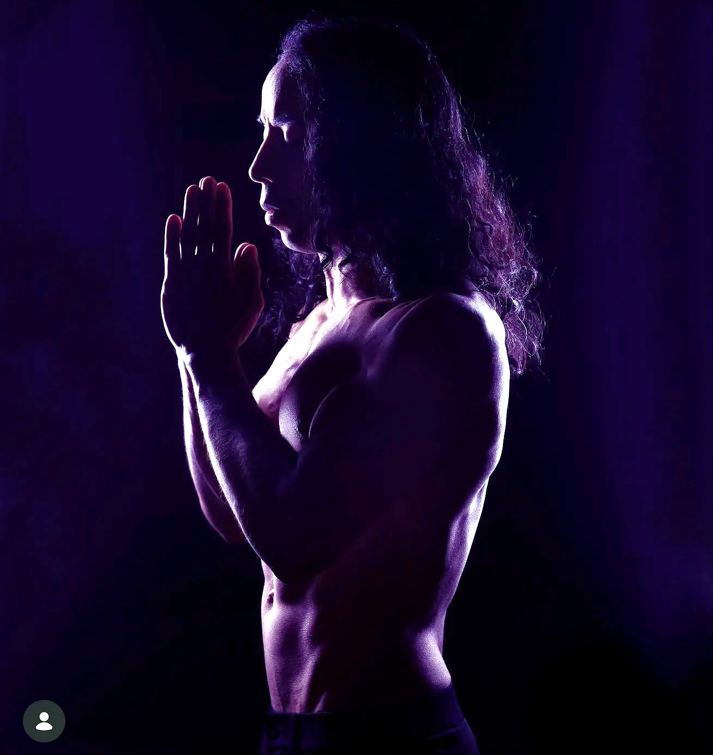 Damien in side profile with hands raised in a prayer pose under purple light